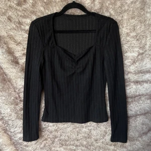 Svart ribbad långärmad topp - Snygg svart långärmad topp med ribbat tyg. Toppen har en unik v-ringad halsringning med lätt rynk framtill. Mjuk och stretchig.