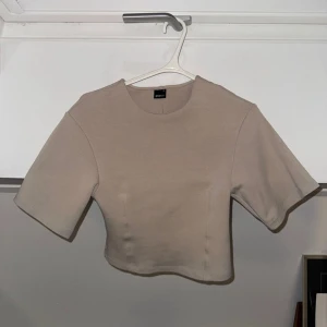 Beige croppad topp från Gina Tricot - Säljer en stilren beige croppad topp från Gina Tricot. Toppen har rund halsringning, korta ärmar och en figurnära passform. Perfekt att matcha med höga jeans eller kjol för en enkel och snygg look.