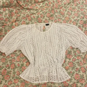 Vit blus med broderade detaljer Gina Tricot - Superfin vit blus från Gina Tricot i storlek 40. Blusen har puffiga korta ärmar, broderade hålmönster och en rund halsringning med knapp i nacken. Midjan är lätt markerad och tyget är luftigt, perfekt för varma dagar.