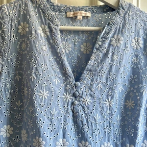 Blå broderad klänning Marie Philippe - Supersöt ljusblå klänning från Marie Philippe med broderade vita blommor, v-ringning och knappar framtill. Klänningen har trekvartsärm och volang i kjolen, perfekt för sommarens alla tillfällen. Tillverkad i bomull för en luftig och skön känsla.