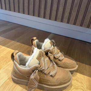 Beige UGG sneakers med fuskpäls - Säljer ett par beige UGG sneakers med breda rutiga snören och mjuk fuskpäls på insidan. Skorna har platt sula, rund tå och detaljer i mocka och syntet. Perfekta för dig som vill ha både stil och komfort under höst och vinter.