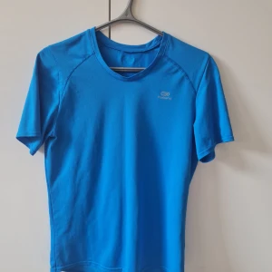 Blå tränings-t-shirt från Decathlon - Säljer en blå tränings-t-shirt från Decathlon med rund halsringning och korta ärmar. Tillverkad i ett lätt och ventilerande syntetmaterial som passar perfekt för träning. Bröstmått 94cm. Kan hämtas i Vallentuna.