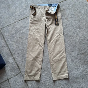 Beiga chinos från Polo Ralph Lauren - Snygga beiga chinos från Polo Ralph Lauren med rak passform och klassisk look. Byxorna har bälteshällor och medföljer ett randigt tygbälte i svart och gult. Perfekta för en clean och stilren outfit.