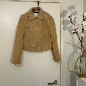Beige mockaskinnjacka från MNG XS - Snygg beige skinnjacka i mocka från MNG med croppad passform och coola detaljer som dragkedjor och knappar. Jackan har klassisk bikerstil med sned dragkedja, axelklaffar och fickor med zip. Perfekt till jeans och t-shirt för en chill look.