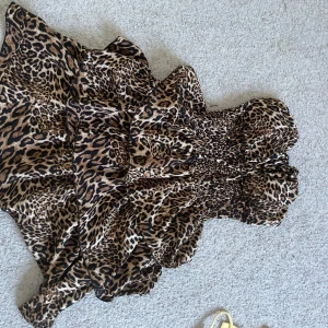 Leopardmönstrad kort klänning - Säljer en snygg leopardmönstrad kort klänning med axelbandslös design och smockad överdel. Klänningen har volanger och knytdetalj framtill, perfekt för dig som vill sticka ut. Färgerna går i brunt, beige och svart. Materialet känns lätt och luftigt.