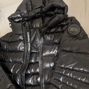 Svart dunjacka från Canada Goose - Säljer en svart, glansig dunjacka från Canada Goose med huva och dragkedja. Jackan har quiltad design, justerbar dragsko och tydlig logotyp på ärmen. Perfekt för kalla dagar och enkel att packa ihop. Klassisk och stilren look. Jackan är helt oanvänd. Jag säljer den pga den är för liten för mig. Storlek S. 