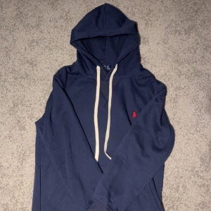 Marinblå hoodie från Polo Ralph Lauren - Snygg marinblå hoodie från Polo Ralph Lauren med vit snörning och klassisk röd logobrodyr på bröstet. Tröjan har huva, långa ärmar och ribbade muddar. Perfekt för chill dagar och enkel att matcha med jeans eller joggers.