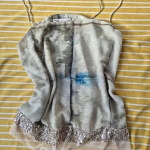 Silkig linne  med paljetter från Zara - Säljer en unik linne från Zara i silkesliknande material med abstrakt mönster i grått, blått och svart. Toppen har smala axelband och en glittrig paljettkant längst ner som ger extra edge. Perfekt för dig som gillar statement-plagg!