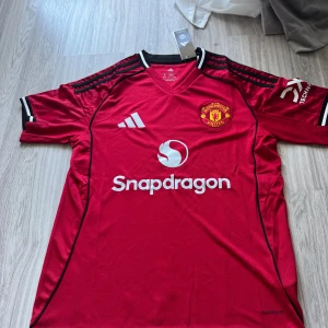 Manchester United hemmaställ  25/26 (Rabatt) - Röd Manchester United fotbollströja från Adidas med klubbmärke, vita loggor och svart-vita detaljer på krage och ärmar. Kortärmad modell i lätt funktionsmaterial med V-ringning och svarta ränder på axlarna. Snapdragon sponsortryck på bröstet.