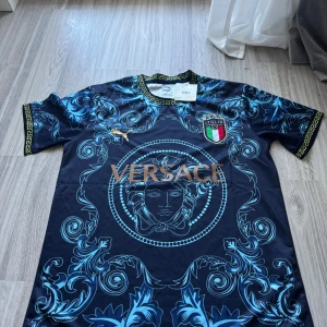 Italien Versace x Puma fotbollströja  - Unik marinblå fotbollströja från Puma i samarbete med Versace och Italien. Tröjan har korta ärmar, guldiga detaljer, klassiskt Versace-mönster i blått och stort Medusa-tryck framtill. Materialet är lätt och andas, perfekt för match eller streetstyle.