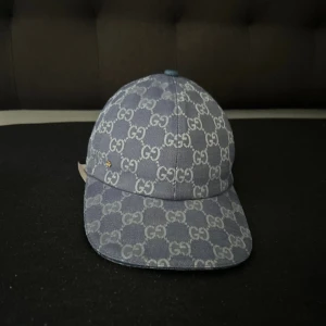 Blå GG keps från Gucci, storlek S - Snygg blå keps från Gucci med klassiskt GG-mönster över hela kepsen. Tillverkad i bomull och har en böjd skärm. Perfekt för dig som vill ha en lyxig och trendig accessoar med tydlig designerstil. Med boxen kostar de extra 