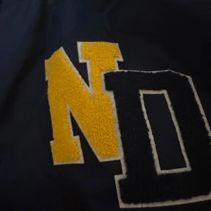 Blå bomberjacka från DNMD med patch - Snygg mörkblå bomberjacka från DNMD med stora gula och svarta bokstavspatchar på bröstet. Jackan har ribbad krage med vita och gula ränder, tryckknappar framtill och meshfoder på insidan. Perfekt för dig som gillar college-vibe och sportig stil.