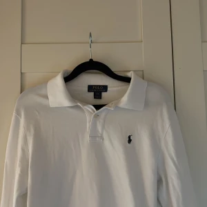 Vit pikétröja från Ralph Lauren - Klassisk vit pikétröja från Polo Ralph Lauren med broderad logga på bröstet. Tröjan har krage, knäppning framtill och är tillverkad i bomull. Perfekt för en clean och stilren look. 