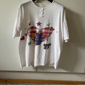 Vit t-shirt med tryck från Veeta - Vit t-shirt från Veeta i bomull med korta ärmar och oversized passform. Framsidan har ett stort tryck med ett rutigt hjärta, blå nalle, stjärnor och rosett. Rund halsringning med knappdetalj framtill. 