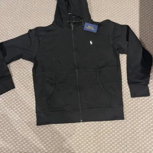 Svart hoodie från Polo Ralph Lauren - Svart hoodie från Polo Ralph Lauren med dragkedja och huva. Klassisk vit logga broderad på bröstet, fickor framtill och snörning i huvan. Tillverkad i mjuk bomull för en skön känsla. Perfekt för en chill och stilren look.