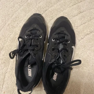 Svarta Nike React sneakers - Svarta Nike React sneakers med vit sula och klassisk vit swoosh på sidan. Skorna har snörning, rund tå och är tillverkade i syntetmaterial och mesh för en sportig look. Sulan har en unik design med svarta detaljer. Perfekta för dig som gillar streetstyle.