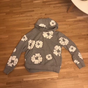 Grå hoodie med blommor från Denim Tears - Grå hoodie från Denim Tears med stora vita blommor tryckta över hela tröjan. Klassisk huva, magficka och ribbade muddar. Perfekt för dig som vill sticka ut med en unik och trendig streetwear-look. Köpt den själv i USA