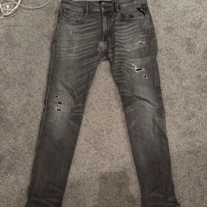 Replay Anbass  - Säljer ett par Replay Anbass jeans i grå tvätt med slitningar och hål på benen. Modellen är slim fit med klassiska fem fickor och dragkedjegylf. Snygga detaljer och cool faded effekt längs benen. Perfekta för dig som gillar en modern och edgy look.