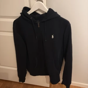 Mörkblå Ralph Lauren hoodie - Mörkblå hoodie från Ralph Lauren, helt utan defekter bortsett från att en metallbricka saknas från ena snöret vilket inte påverkar helhetsintrycket överhuvudtaget. Skick Mycket bra. Nypris ca 2300kr mitt pris 589kr