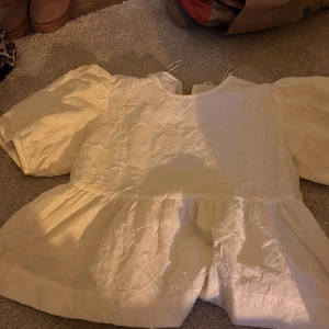 Vit blus med puffärm från Gina Tricot - Supersöt vit blus från Gina Tricot i storlek 170. Blusen har puffiga korta ärmar, rund hals och volang nedtill. Materialet är glansigt och har en lätt skrynklig struktur som ger cool känsla. Perfekt för dig som gillar romantisk och trendig stil.