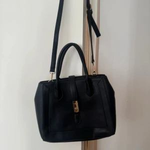 Svart handväska med gulddetaljer - Stilren svart handväska i skinnimitation med både handtag och justerbar axelrem. Väskan har guldfärgade metalldetaljer, lås framtill och fötter undertill. Insidan har dragkedjefack och flera praktiska fack för dina grejer. Perfekt storlek för vardagsprylar.