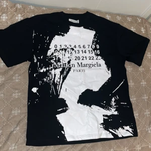 Maison Margiela svart t-shirt - Maison Margiela t-shirt med stor vit abstrakt print och ikonisk sifferlogga framtill. Klassisk passform med rund hals och korta ärmar. Tillverkad 100% bomull för en skön känsla. Tveka inte vid frågor eller funderingar!!