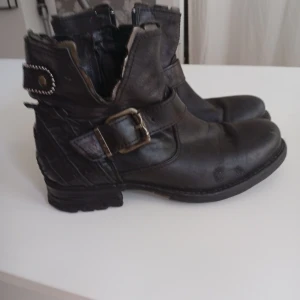 Svarta boots från Replay med spänne - Säljer ett par svarta boots från Replay i skinn med coola detaljer som metallspänne och nitar bak. Snyggt mönstrad bakdel och dragkedja på sidan för enkel påtagning. Rund tå och platt sula, perfekt för dig som gillar en edgy stil.