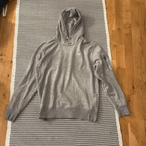 Grå hoodie från Alpha Industries - Säljer en stilren grå hoodie från Alpha Industries i storlek S. Tröjan har huva med snygga metallöglor, lång ärm med ficka på ena ärmen och diskret logga på bröstet. Tillverkad i mjuk bomullsmix, perfekt för chill dagar.