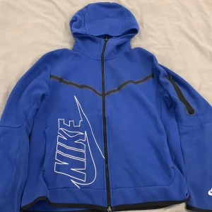 Blå Nike hoodiejacka med dragkedja - Blå hoodiejacka från Nike med hel dragkedja, svart detalj längs bröstet och ärmficka med dragkedja. Stor Nike-logga i vitt på framsidan. Jackan har huva och är långärmad, perfekt för en sportig look.