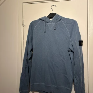 Blå hoodie från Stone Island - Säljer en snygg blå äkta hoodie från Stone Island med klassisk patch på vänster ärm. Tröjan har huva med snören. Väldigt bra skick utan skador eller liknande, STORLEK Medium 