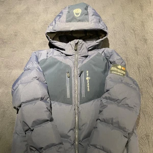 Sail Racing jacka - Snygg grå dunjacka från Sail Racing med huva och två bröstfickor med dragkedja. Jackan har logga på bröstet och ärmen, samt coola paneler i mörkare grått. Perfekt för kalla dagar och riktigt skön passform.