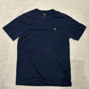 Mörkblå t-shirt från Polo Ralph Lauren - Klassisk mörkblå t-shirt från Polo Ralph Lauren med broderad vit logga på bröstet. T-shirten har rund halsringning och korta ärmar. Tillverkad i mjuk bomull för en skön och avslappnad känsla. Den är XL i barn storlek vilket motsvarar S i man storlek.