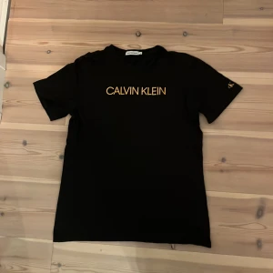 Svart Calvin Klein Jeans t-shirt - Svart t-shirt från Calvin Klein Jeans med gul logga framtill och gul detalj med texten 'JEANS' bak. Klassisk passform, rund hals och korta ärmar. Tillverkad i mjuk bomull för en skön känsla.