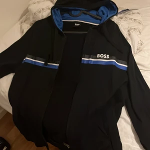 Svart kofta från BOSS med huva - Svart vindjacka från BOSS med huva och blått foder. Jackan har vita och blå ränder över bröstet, dragkedja framtill och BOSS-logga på vänster sida. Snygg och sportig design med stilrena detaljer, perfekt för dig som gillar streetwear.