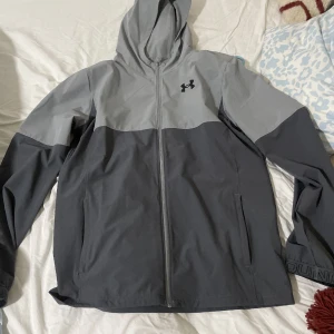 Grå vindjacka från Under Armour - Snygg tvåtonad grå vindjacka från Under Armour med huva och hel dragkedja framtill. Jackan har långärmade ärmar, två sidofickor och logga på bröstet. Perfekt för blåsiga dagar och sportiga outfits.