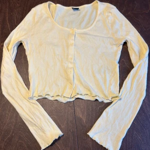 Ljusgul ribbad topp från Gina Tricot - Supersöt ljusgul ribbad topp från Gina Tricot med långa ärmar och knappar framtill. Modellen är croppad med vågig kant nertill och har en mjuk, stretchig känsla. 
