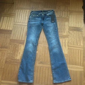 True Religion Becca Bootcut Jeans W26 - Oanvända blå jeans från True Religion, modell Becca, midrise bootcut flap. Prislappar finns fortfarande kvar. Jeansen har klassisk femficksdesign, coola bakfickor med lock och gul kontrastsöm samt silverfärgade knappar. Tillverkade i jeansmaterial med en lätt tvättad look och markerade sömmar. Gula och vita sömmar.