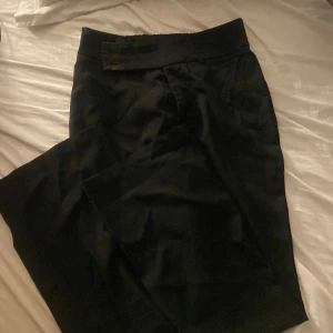 Svarta kostymbyxor med knappar - Snygga svarta kostymbyxor med klassisk passform. Byxorna har två knappar i midjan och hake för extra säker stängning men den är tyvärr lös men kan lätt sys fast, men den funkar lika bra utan. De är raka i modellen och har diskreta fickor bak. Perfekta för en stilren look.