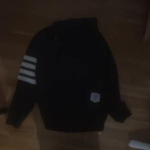 Svart sweatshirt från Thom Browne - Svart sweatshirt från Thom Browne med vita ränder på ena ärmen och en vit patch med märkets logga framtill. Tröjan har en klassisk rund hals och lång ärm, perfekt för dig som gillar stilrena detaljer och streetwear-vibe.