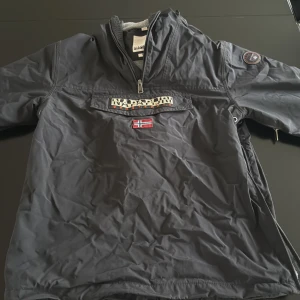 Svart vindjacka från Napapijri - Säljer en svart vindjacka från Napapijri med halv dragkedja och stor logga på bröstet. Jackan har norsk flagga framtill, huva och praktisk ficka på magen. Perfekt för blåsiga dagar och streetwear-stil.