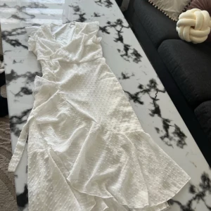 Vit omlottklänning med volang - Supersöt lång vit klänning med omlott-design och volangdetaljer. Klänningen har prickigt mönster i tyget och knytband i midjan för en snygg passform. Perfekt för sommarens alla tillfällen, med lätt och luftig känsla.