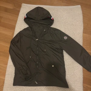 Mörk grön Moncler vindjacka med huva - Snygg mörkgrön vindjacka från Moncler med huva och dragsko, klassisk logga på ärmen och dragkedja framtill. Jackan har diskreta fickor med knapp och coola röd-vita detaljer på huvan. Perfekt för dig som gillar stilren och sportig look.