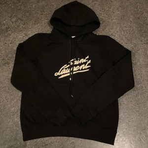 Saint Laurent tröja - Säljer en snygg svart hoodie från Saint Laurent med deras ikoniska logga i vitt på bröstet. Perfekt för både vardag och fest! Den har en klassisk passform med justerbar huva och ribbade muddar. Storlek S men passar även M. Superbekväm och stilren, ett måste i garderoben!