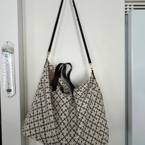 Mönstrad shoppingväska By Malene Birger - Stor shoppingväska från By Malene Birger i beige och svart med deras ikoniska grafiska mönster. Väskan har svarta handtag och en lång axelrem för enkel användning. Tillverkad i slitstark polyester, perfekt för dig som vill ha plats för allt.