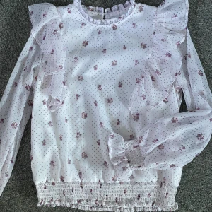 Vit blommig blus med volang - Supersöt vit blus med små lila blommor och diskreta prickar. Blusen har långa ärmar, volangdetaljer på axlarna och smockad nederkant samt ärmslut. Sitter som xs❣️