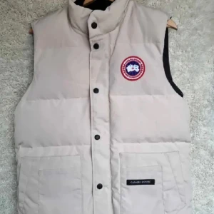 Beige dunväst från Canada Goose - Snygg beige dunväst från Canada Goose med klassisk logga på bröstet. Västen har hög krage, stora framfickor och stängs med både dragkedja och knappar. håller dig varm under kyliga dagar.