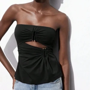 Svart tubtopp med cut-out och snörning - Snygg svart tubtopp med cool cut-out detalj framtill och snörning med små pärlor. Toppen är axelbandslös och har en unik design som sticker ut. Perfekt att matcha med jeans eller kjol för en trendig look.