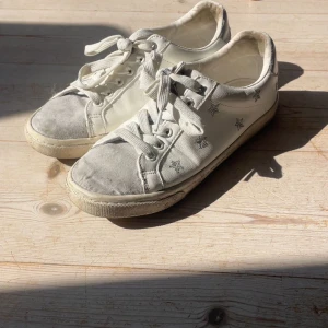 Vita sneakers med stjärnmönster - Snygga vita sneakers med silverfärgade stjärnor broderade på sidorna. Skorna har rund tå, platt sula och klassisk snörning. Tåpartiet är i ljusgrå mocka och hälen har en diskret silverdetalj. Perfekta för dig som gillar en clean look med lite extra detaljer.