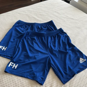Blå sportshorts från Adidas - Säljer två par blå sportshorts från Adidas med vitt tryck 'FH' och Adidas-logga. Kan säljas i par men även själva oxå! En har kvar lappen och en inte. Jättesköna och luftiga perfekt till träning som fotboll.!!