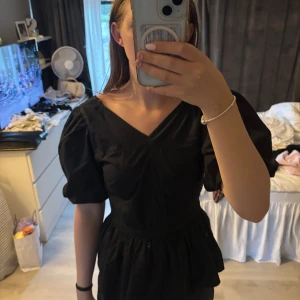 Svart blus med puffärmar från C - Supersnygg svart blus med v-ringning, puffiga korta ärmar och volang nedtill. Har också en fin liten rosett i ryggen som går att knyta, är i super fint skick
